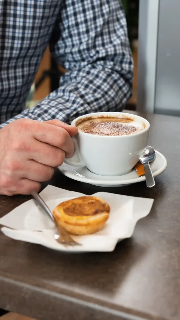 En hånd holder en kop kaffe ved siden af en pastel de nata.